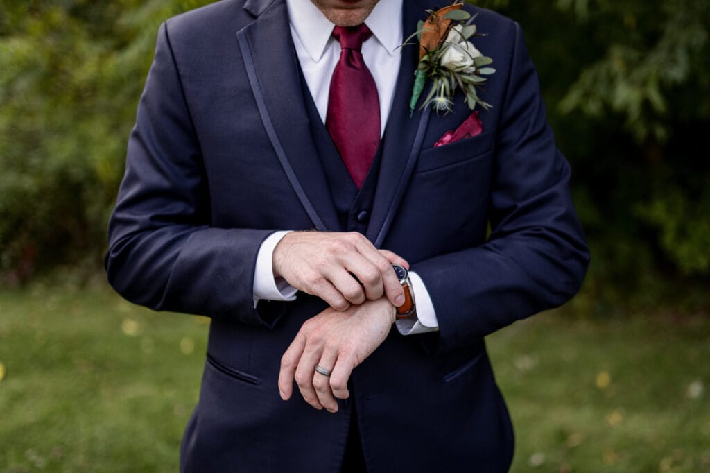 Navy Blue Suit Man in a navy suit adjusts his watch, with a boutonniere and red tie, standing in a green outdoor setting.