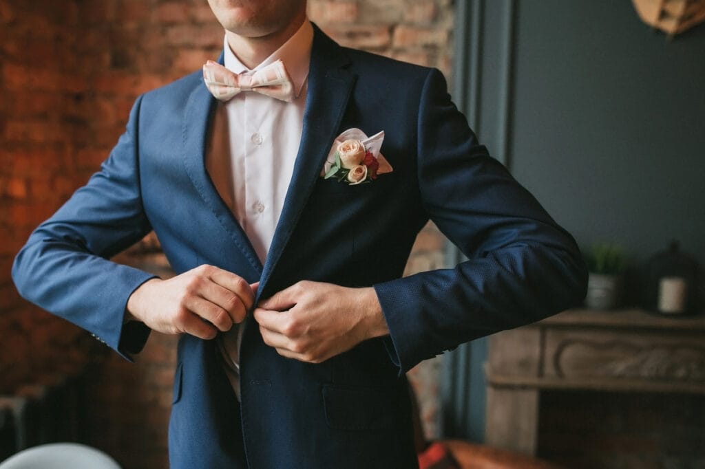 Wedding Suits for Men A person in a blue suit adjusts their jacket in a warmly lit room with a brick wall background.